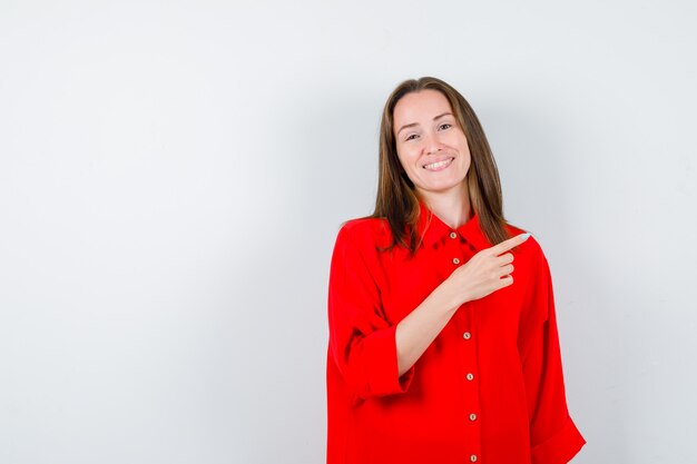 Young woman pointing right in red blouse and looking cheery , front view.