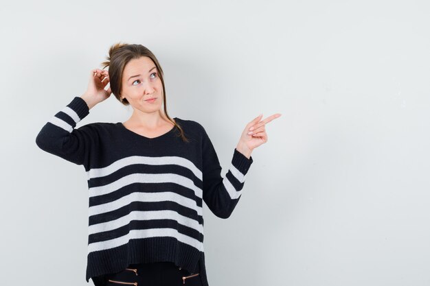 Young woman pointing right and scratching head in striped knitwear and black pants and looking pensive