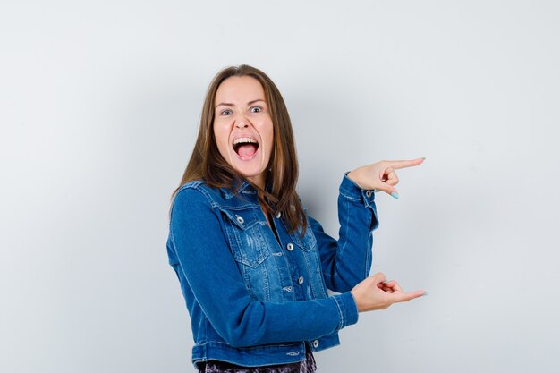 Young woman pointing to the right side, opening mouth in denim jacket, dress and looking furious. front view.