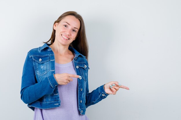 Young woman pointing right in t-shirt, jacket and looking cheery , front view.