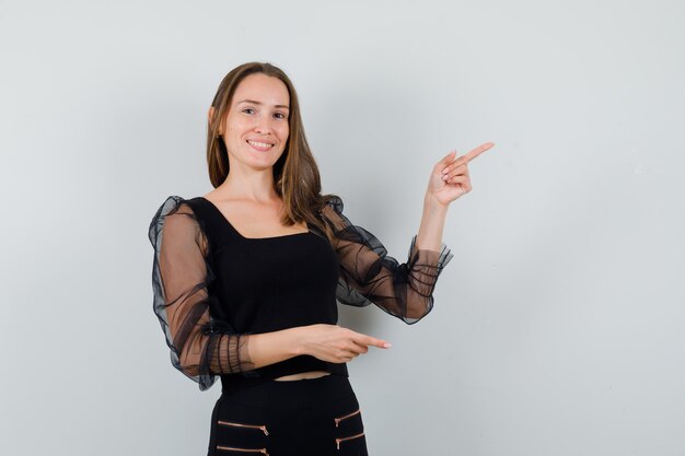 Young woman pointing right with both index fingers in black blouse and black pants and looking optimistic 