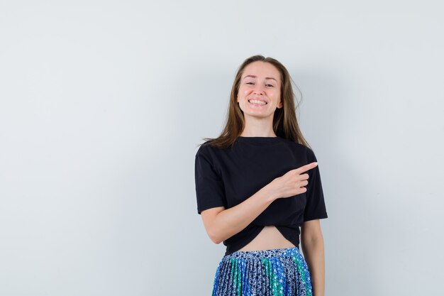 Young woman pointing right with index finger in black t-shirt and blue skirt and looking happy