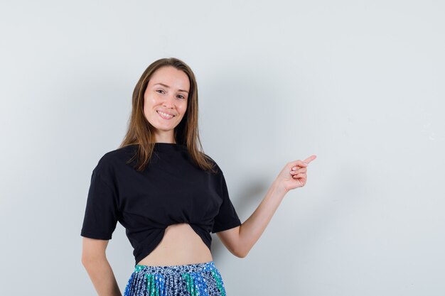 Young woman pointing right with index finger in black t-shirt and blue skirt and looking happy