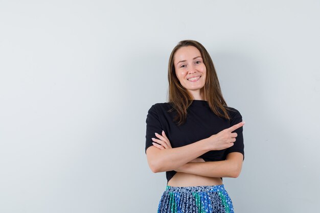 Young woman pointing right with index finger in black t-shirt and blue skirt and looking happy