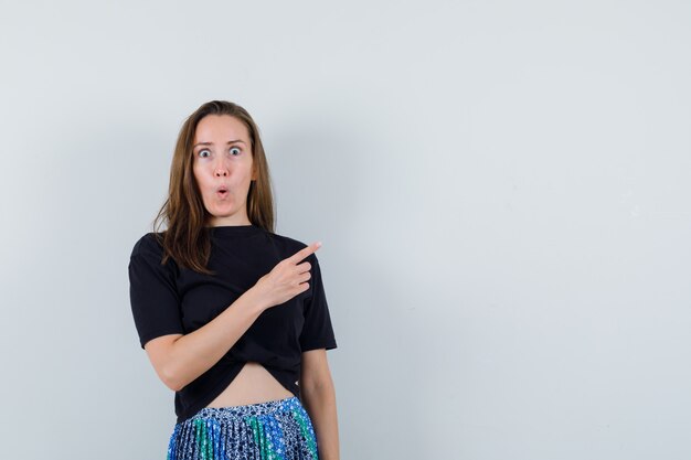 Young woman pointing right with index finger in black t-shirt and blue skirt and looking surprised