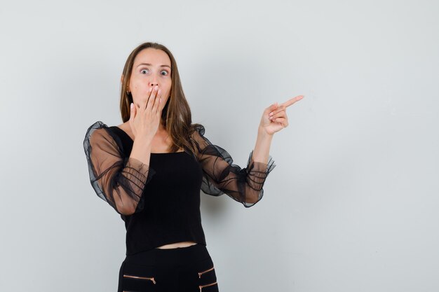 Young woman pointing right with index finger and covering mouth with hand in black blouse and black pants and looking surprised. front view.