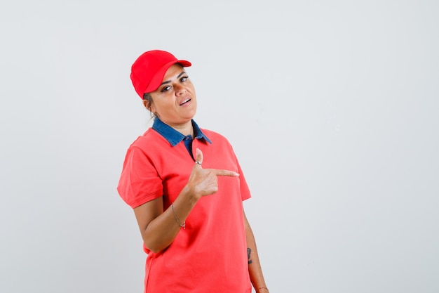 Young woman pointing right with index finger in red shirt and cap and looking pretty. front view.