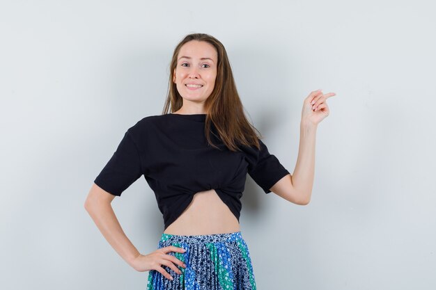 Young woman pointing right with index finger while holding hand on waist in black t-shirt and blue skirt and looking happy