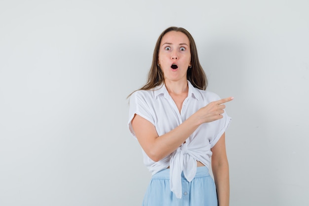 Young woman pointing right with index finger in white blouse and light blue skirt and looking surprised