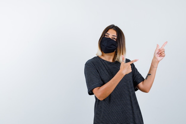 Young woman pointing right with index fingers in black dress, black mask and looking serious , front view.