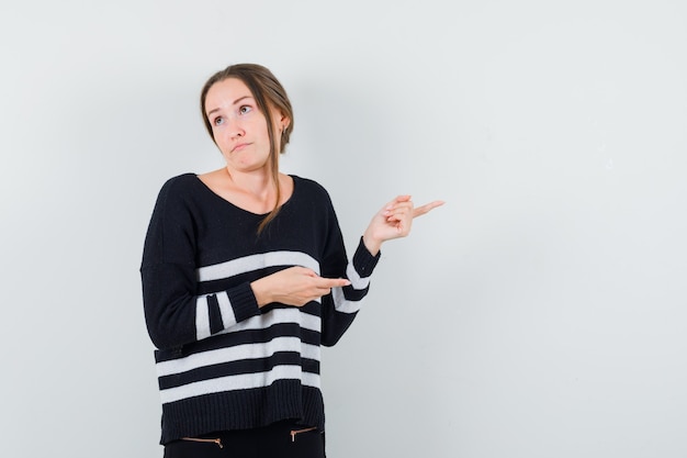 Giovane donna che punta a destra con il dito indice e distoglie lo sguardo in maglieria a righe e pantaloni neri e sembra tormentata