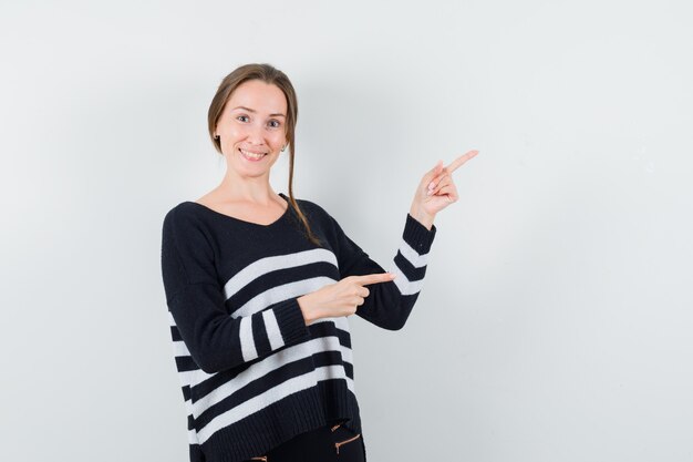 Young woman pointing right with index fingers in striped knitwear and black pants and looking happy