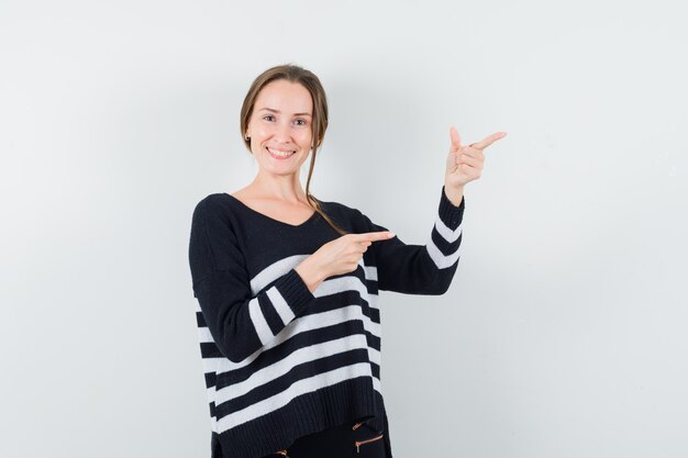 Young woman pointing right with index fingers in striped knitwear and black pants and looking happy