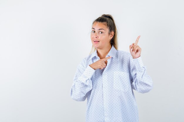 Молодая женщина указывает вправо указательными пальцами в белой рубашке и выглядит счастливой