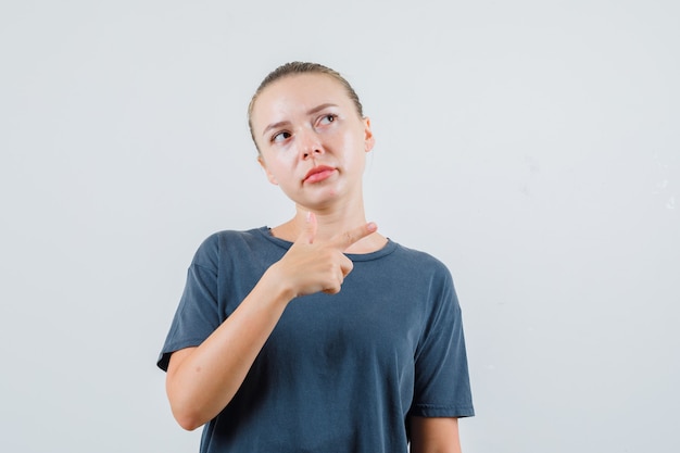 Young woman pointing to side in grey t-shirt and looking confused