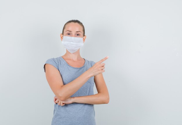 Young woman pointing to side in grey t-shirt, mask , front view.