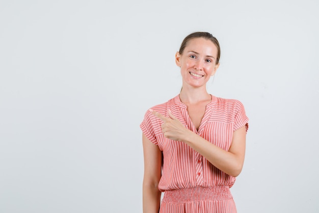 Young woman pointing to side in striped dress and looking cheerful , front view.