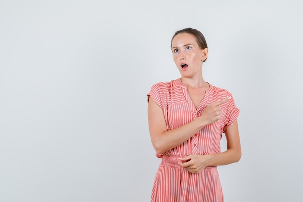 Young woman pointing to side in striped dress and looking surprised , front view.