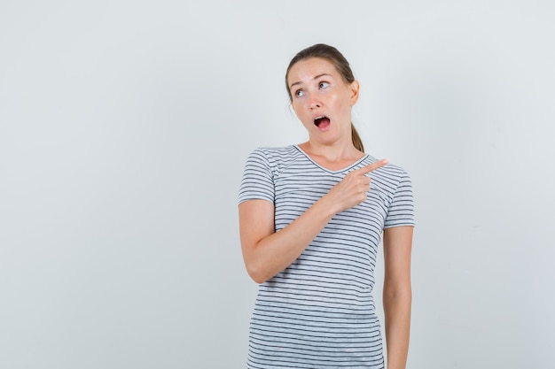 Young woman pointing to side in t-shirt and looking confused. front view.