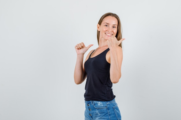 Young woman pointing thumbs back in singlet, shorts and looking cheery .