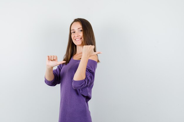 紫色のシャツに親指を向けて幸せそうに見える若い女性。正面図。