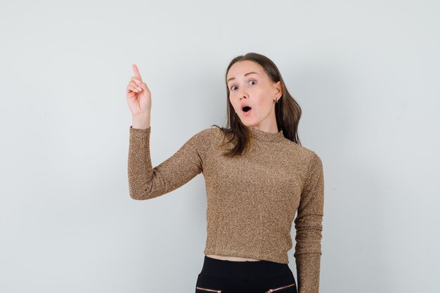 Young woman pointing up in golden blouse and looking surprised. front view.