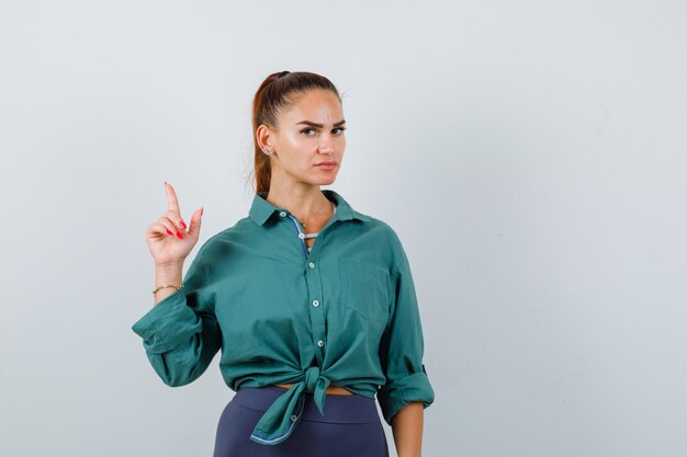 Young woman pointing up in green shirt and looking careful , front view.