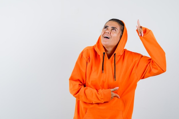 Young woman pointing up and holding hand on belly in orange hoodie and looking happy