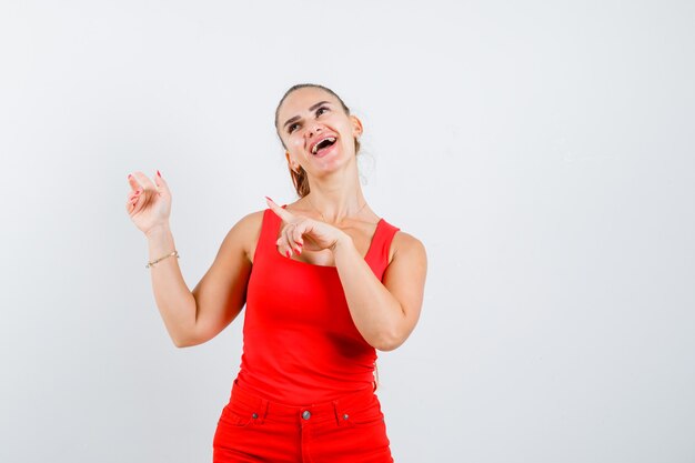 Young woman pointing up in red tank top, pants and looking cheerful , front view.