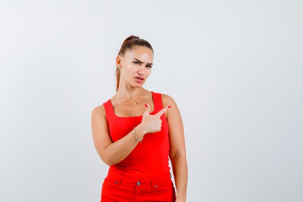 Young woman pointing up in red tank top, pants and looking thoughtful , front view.
