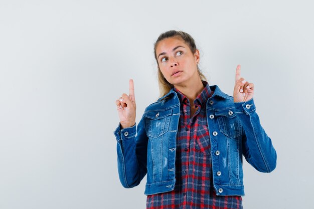 Young woman pointing up in shirt, jacket and looking pensive. front view.