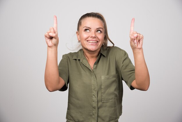 Young woman pointing at up side on gray wall.