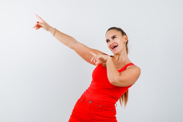 Free photo young woman pointing up, standing sideways in red tank top, pants and looking cheerful .