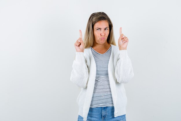 Young woman pointing up in t-shirt, jacket and looking displeased , front view.