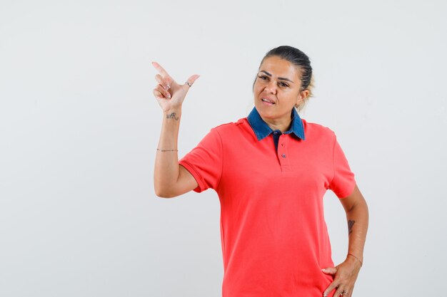 Young woman pointing up with index finger and holding hand on waist in red t-shirt and looking displeased. front view.