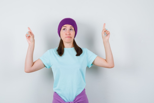 Молодая женщина указывая вверх указательными пальцами в синей футболке, фиолетовой шапочке и выглядит мило, вид спереди.