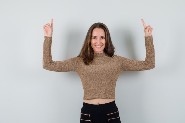 Young woman pointing up with index fingers in gold gilded sweater and black pants and looking happy