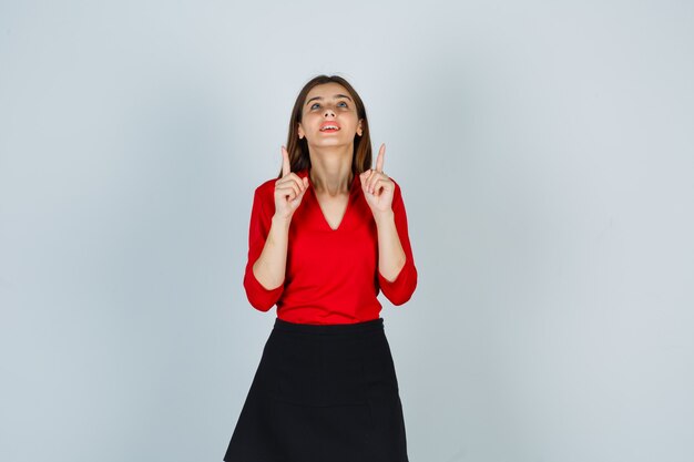 Young woman pointing up with index fingers in red blouse, black skirt and looking happy