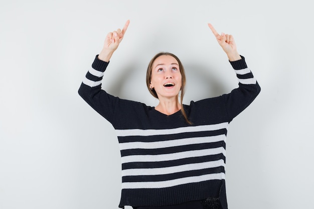 Young woman pointing up with index fingers in striped knitwear and black pants and looking surprised