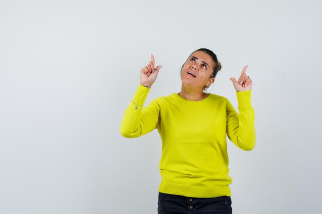 Young woman pointing up with index fingers in yellow sweater and black pants and looking focused