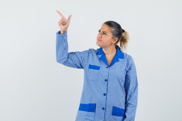 Молодая женщина, указывая на верхнюю левую сторону указательным пальцем в синей пижамной рубашке в клетку, выглядит серьезной. передний план.