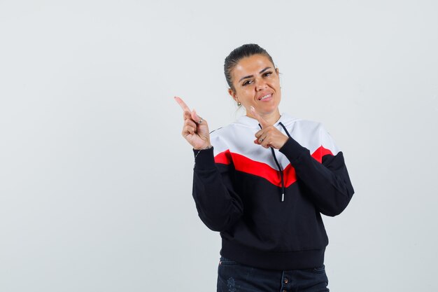 Young woman pointing upper left with index fingers in sweater and black jeans and looking happy , front view.