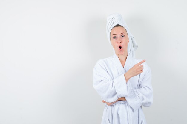 Молодая женщина, указывая на верхний правый угол в белом халате, полотенце и глядя изумленно, вид спереди.