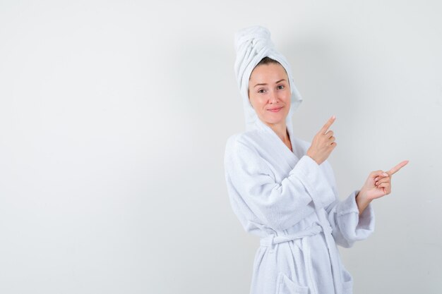 Young woman pointing at upper right corner in white bathrobe, towel and looking merry. front view.