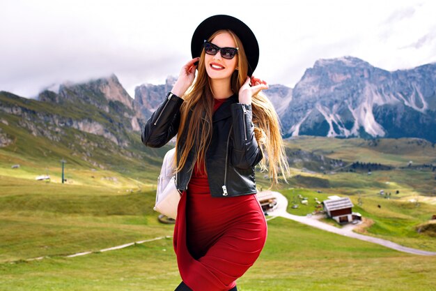 알프스 산맥에서 포즈를 취하는 젊은 여성, 드레스, 가죽 재킷, 선글라스 및 배낭 착용