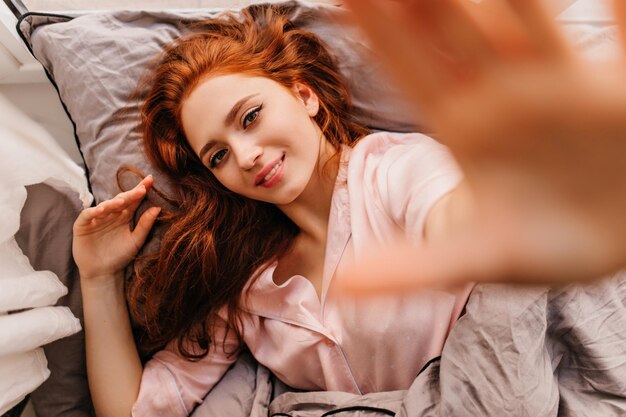 Молодая женщина позирует в постели с искренней улыбкой Внутренний снимок смеющейся рыжей девушки, делающей селфи утром