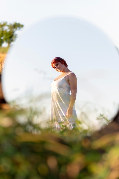Молодая женщина уверенно позирует на открытом воздухе с круглым зеркалом