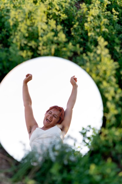 Free photo young woman posing confidently outdoors using a round mirror