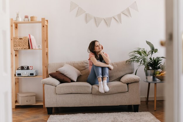 Young woman posing dreamily, sitting on sofa in her bright apartment