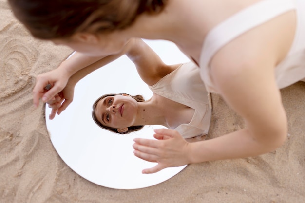 Молодая женщина позирует на открытом воздухе на пляже, используя круглое зеркало